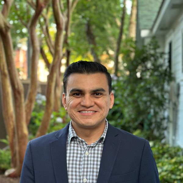 Man wearing a checkered shirt and blazer smiling outdoors.