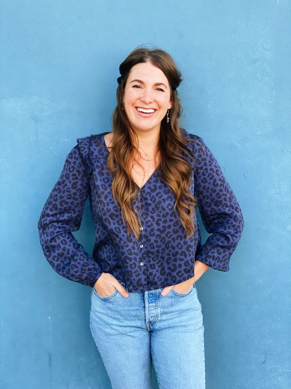 Smiling woman in a blue patterned blouse and jeans against a blue wall.