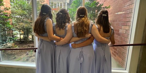 Four women in matching lavender dresses stand arm-in-arm facing a large window.