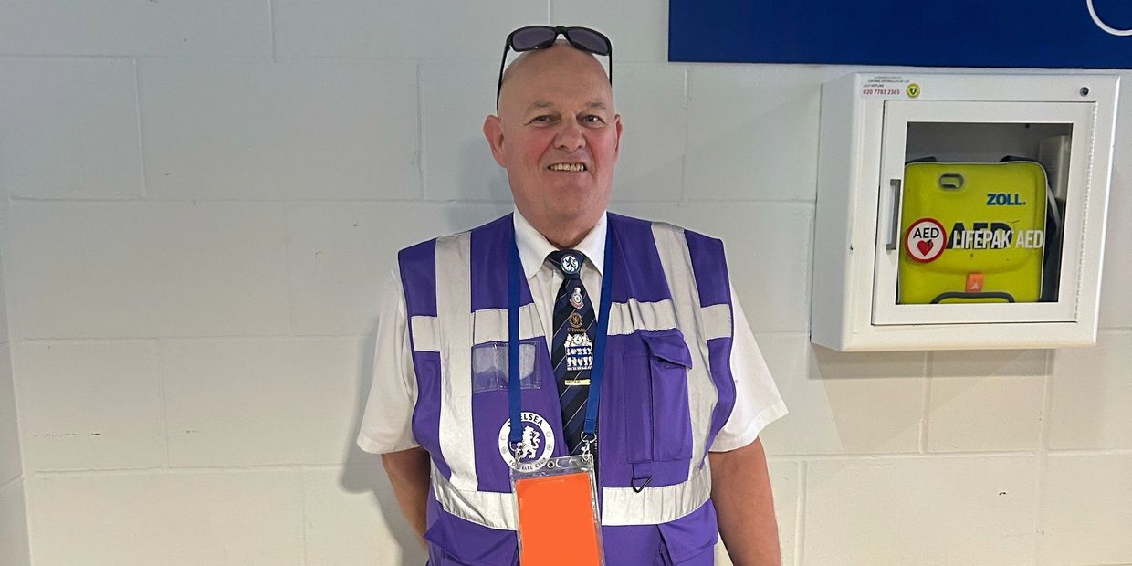 A male steward stands in front of a white wall in a purple visibility bib.