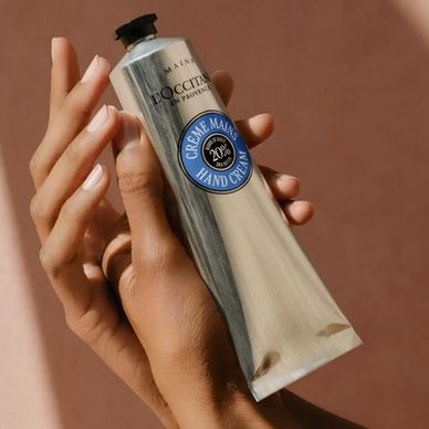 Hand holding a silver tube of L'Occitane hand cream against a brown background.
