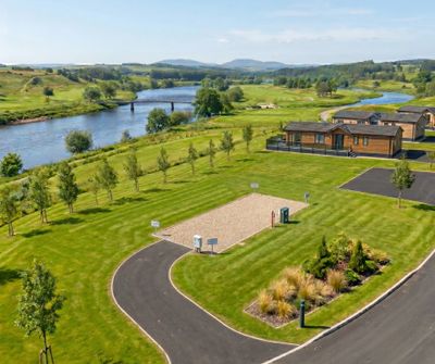 Scenic riverside park with greenery, paths, and small wooden buildings under a clear sky.