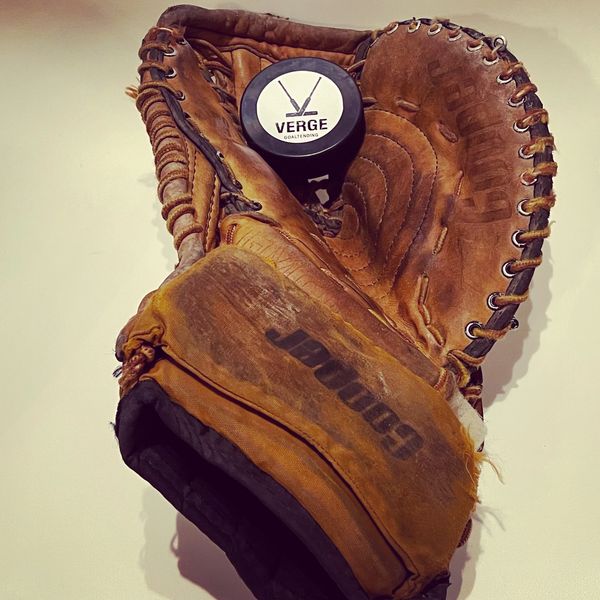 Worn baseball glove holding a hockey puck labeled Verge Goaltending.