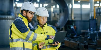 Two engineers in safety gear discussing plans on a laptop in an industrial setting.