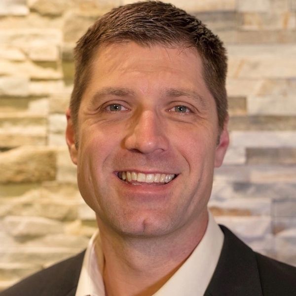 Smiling man in a suit jacket with a stone wall background.