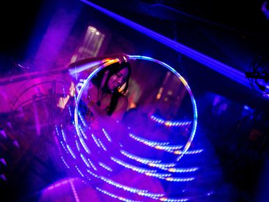 A woman performs with a glowing LED hula hoop in a dark, colorful setting.