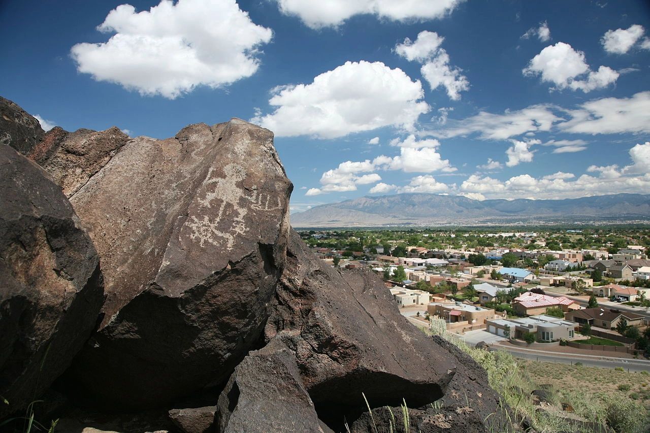 Petroglyphs on rocks overlooking a suburban area with mountains and clouds.