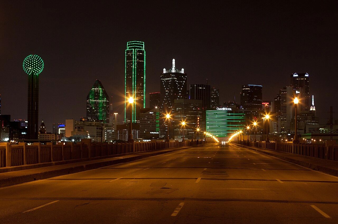 Night view of a city skyline with green-lit buildings and an empty street.