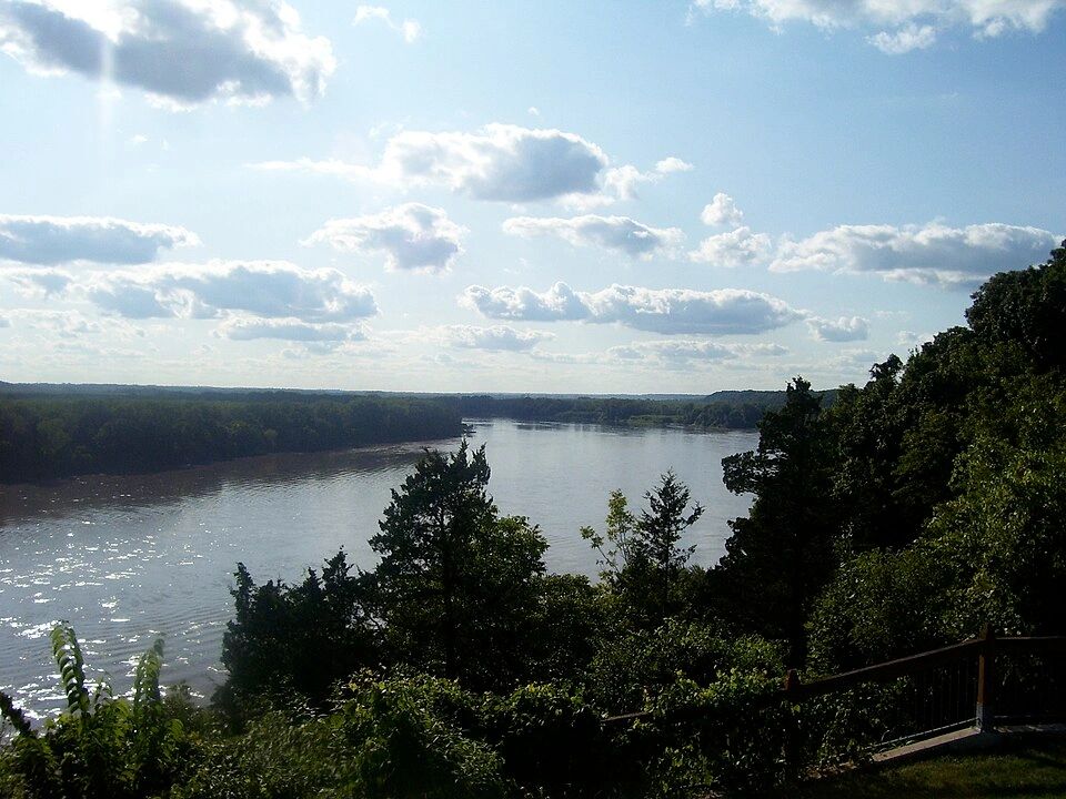 A serene river flowing through a lush forest under a partly cloudy sky.