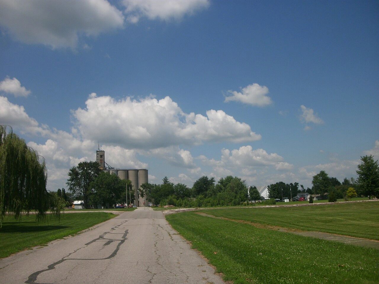 A rural road leading to silos under a bright blue sky with scattered clouds.