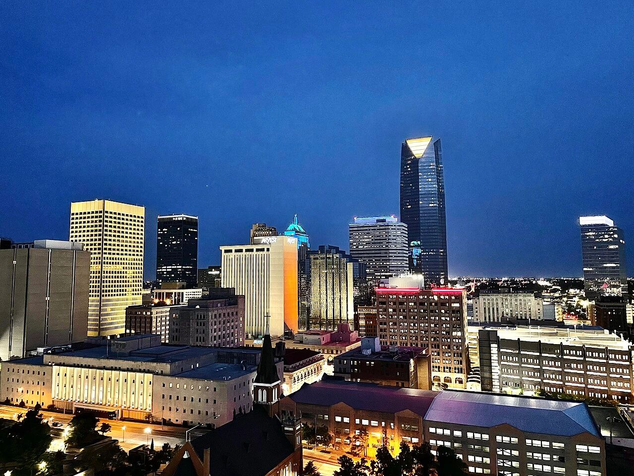 Nighttime cityscape with illuminated skyscrapers under a clear blue sky.