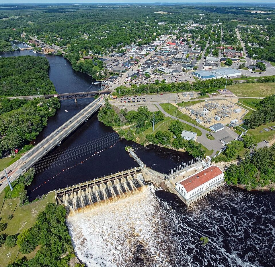 Aerial view of a dam with water flowing over, bridges, and a nearby town surrounded by greenery.
