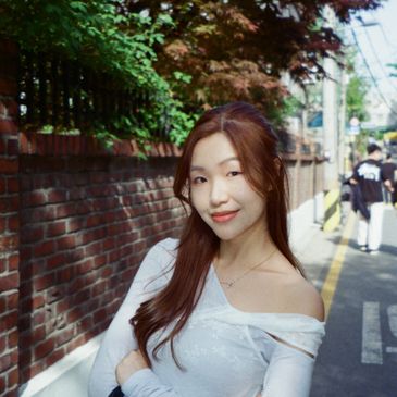Young woman posing outdoors with a brick wall and greenery in the background.