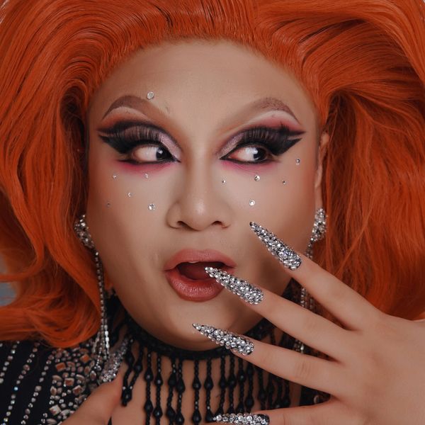Person with vibrant orange hair and dramatic makeup, adorned with rhinestone nails and facial gems.