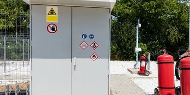 Electrical cabinet with multiple safety warning signs outdoors near fire extinguishers.