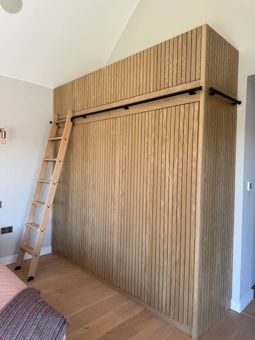 Tall wooden wardrobe with vertical grooves and a ladder for upper access.