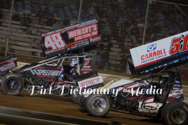 Two sprint cars racing on a dirt track at night.