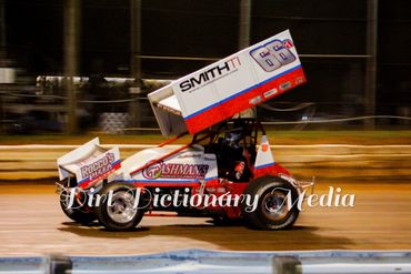 Red and white sprint car racing on dirt track at night.