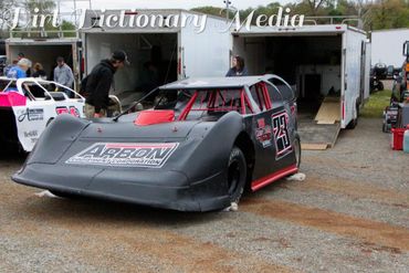 A black and red dirt late model race car parked outside a trailer.