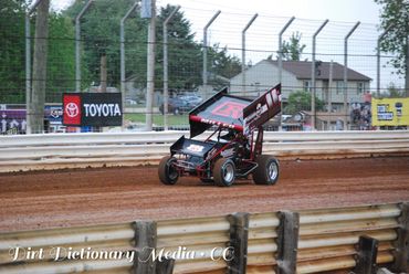 A sprint car races on a dirt track under overcast skies.
