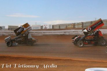 Two sprint cars racing on a dirt track under clear skies.