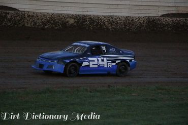 Blue stock car number 24R racing on a dirt track at dusk.