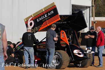 Crew members prepare a sprint car with number 6 for racing at a dirt track.