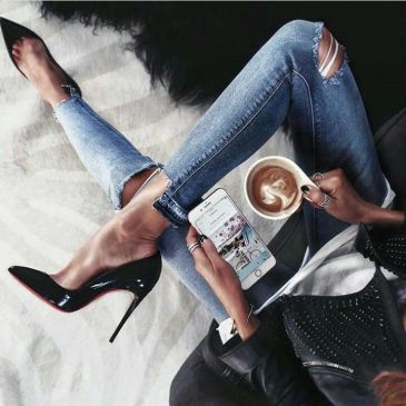 Woman in ripped jeans with high heels, holding phone and coffee.