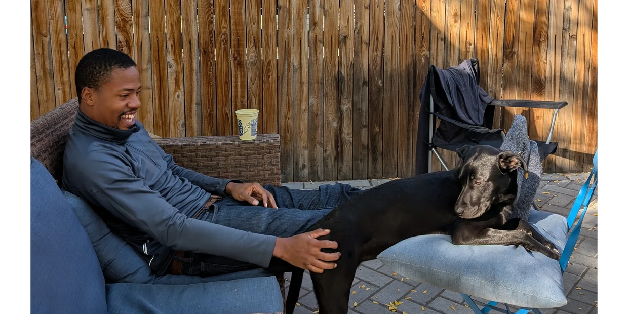 Man sitting on a couch petting a black dog resting on a cushion.