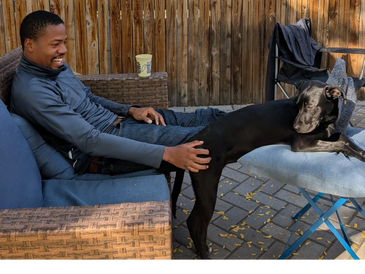 Man sitting on a couch petting a black dog resting on a cushion.