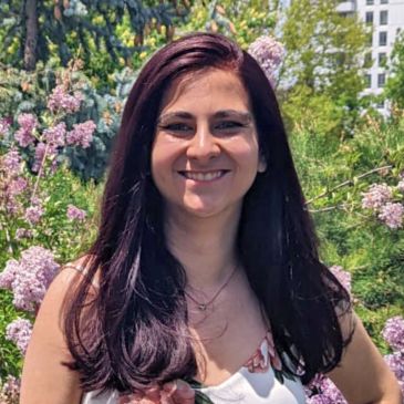 Smiling woman in floral dress stands amidst blooming lilacs on a sunny day.