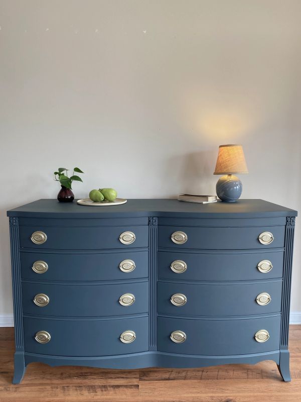 Elegant blue dresser with brass handles, decorated with a lamp, book, plant, and fruit plate.