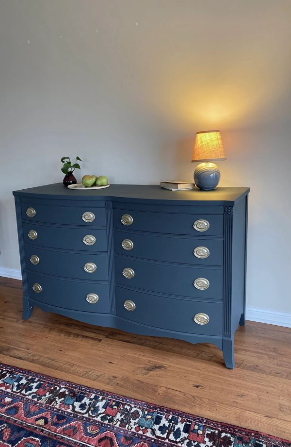 Elegant blue dresser with brass knobs, lamp, and fruit on top in a cozy room.