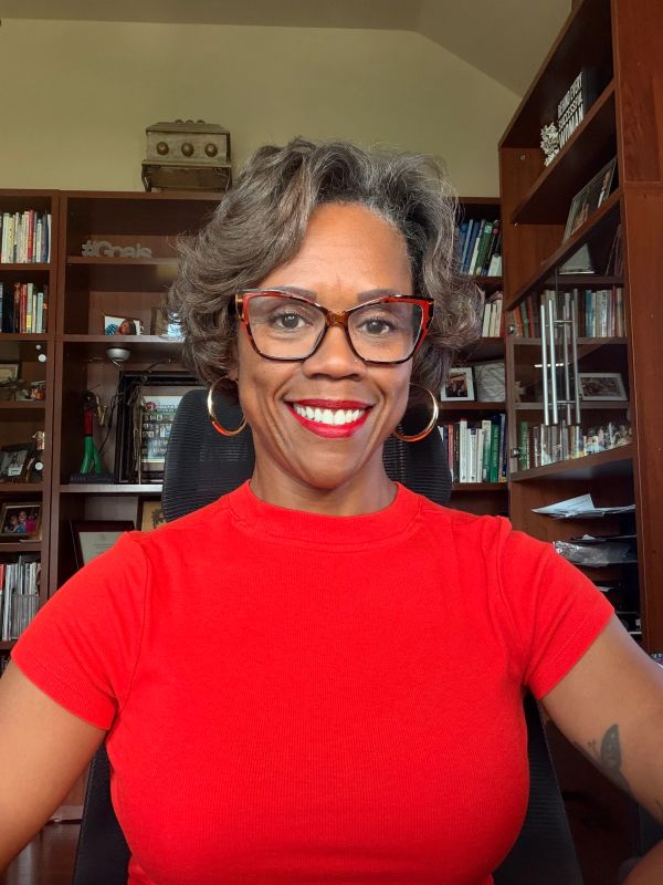 Smiling woman in red shirt with glasses sitting in front of bookshelves.