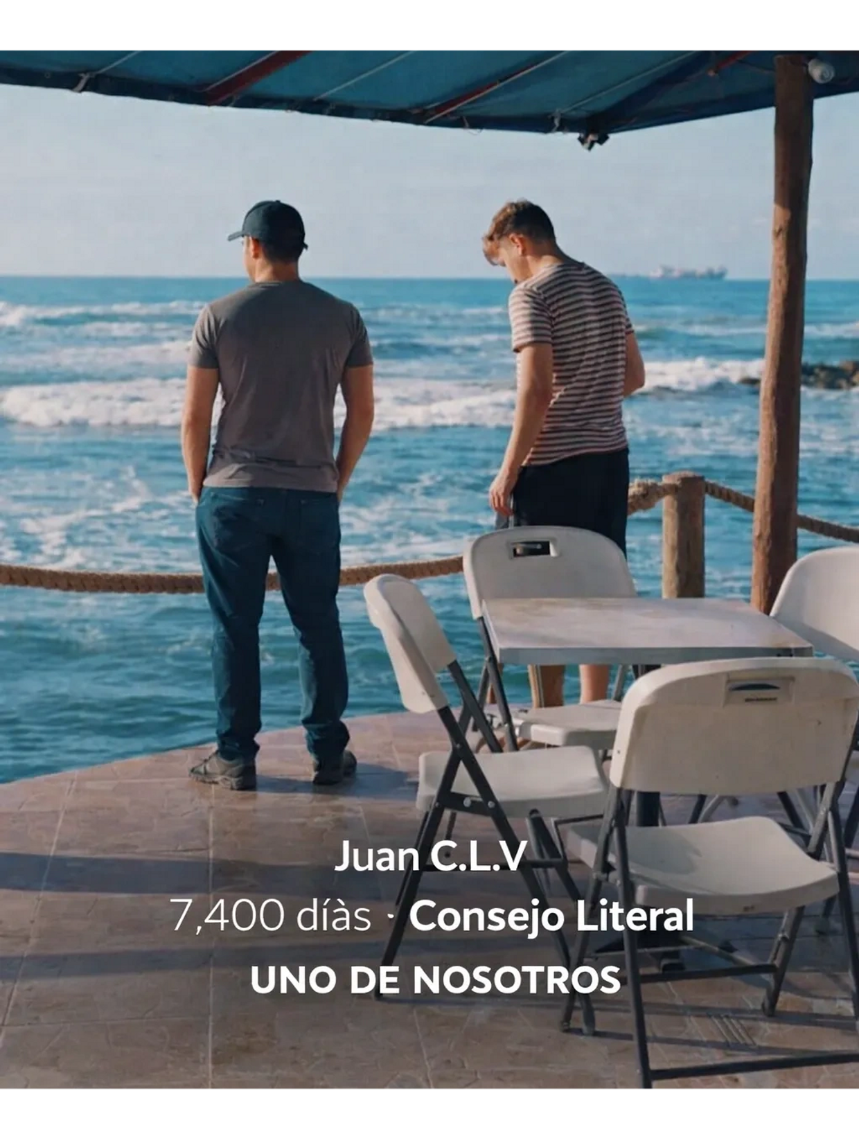 Two men stand by the ocean under a shaded area with empty chairs and a table.