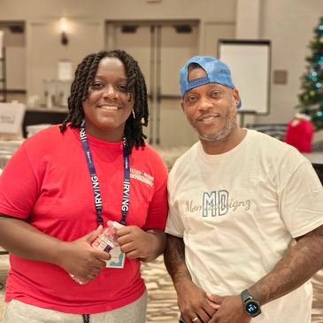 Two people smiling indoors, one in a red shirt, one in a white shirt and blue cap.