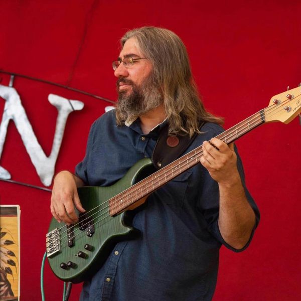 Man with long hair playing a green electric bass guitar against a red background.
