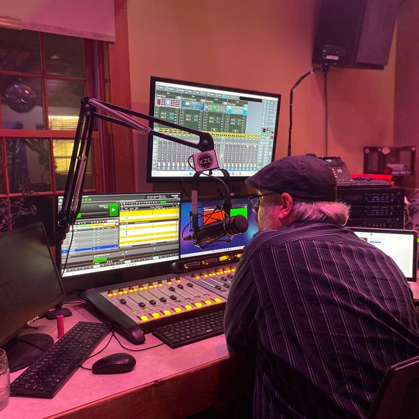 A man operates audio equipment in a radio station studio with multiple monitors and a microphone.