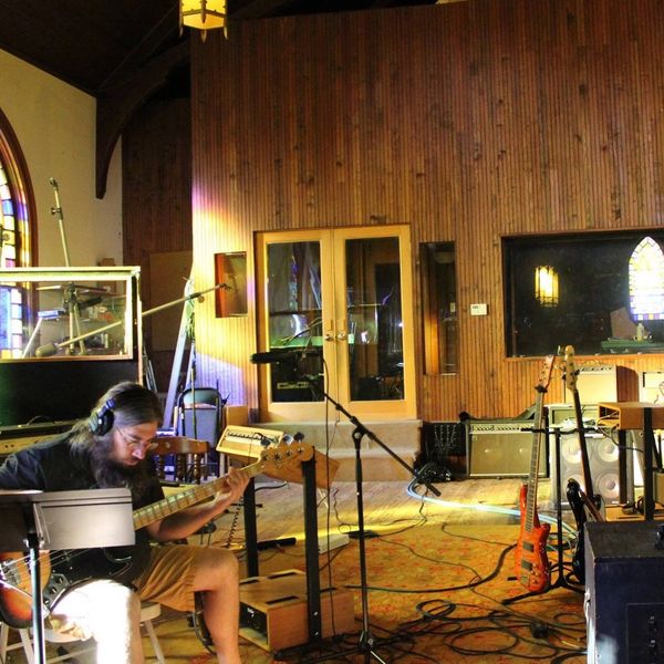 Musician plays bass guitar in a cozy recording studio with wooden walls.