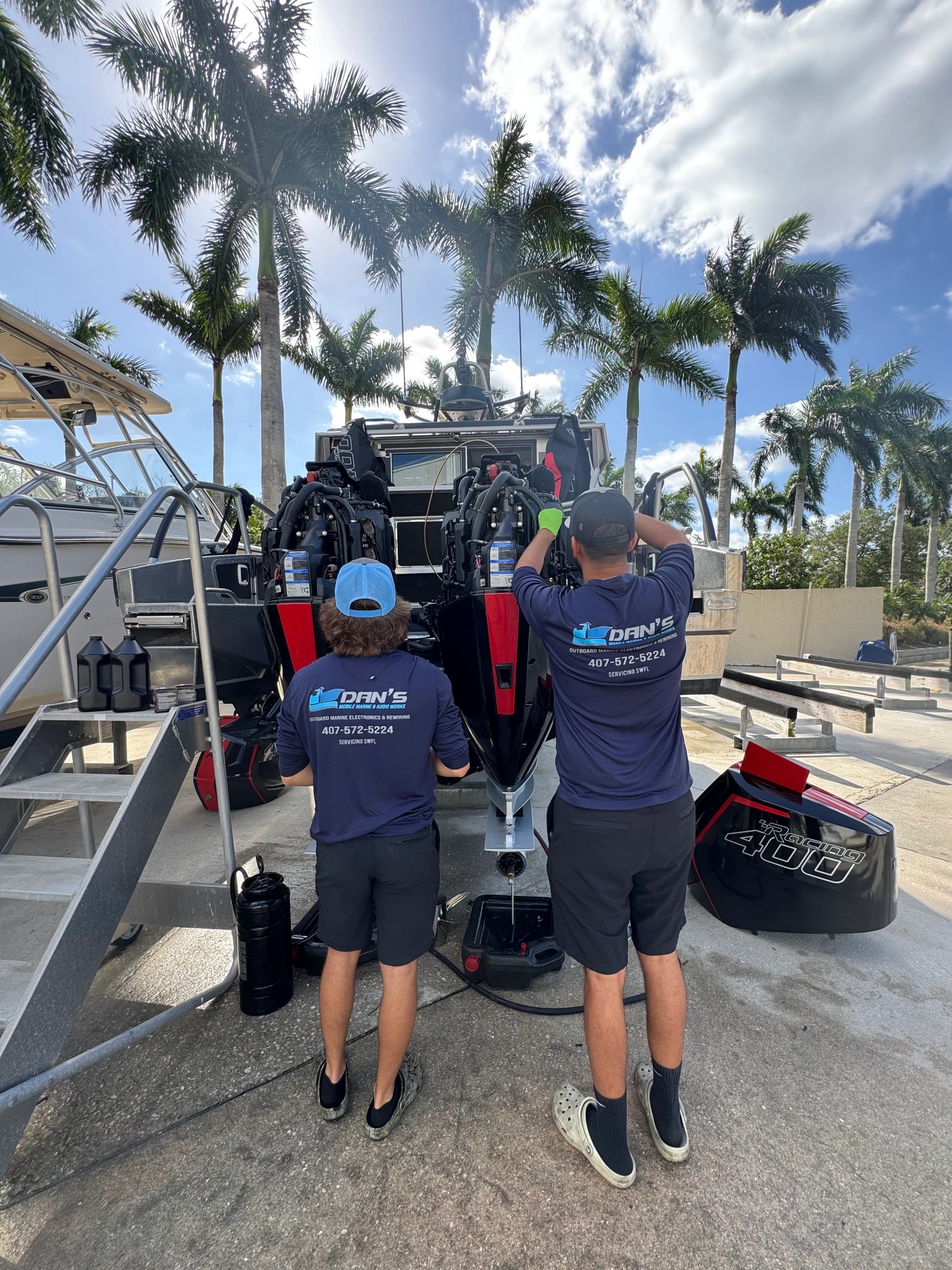 Two men servicing boat engines outdoors under palm trees.