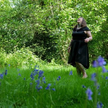 Woman in black dress laughing in a green forest with purple flowers.