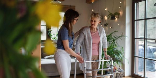 A young woman assists an elderly woman using a walker indoors.