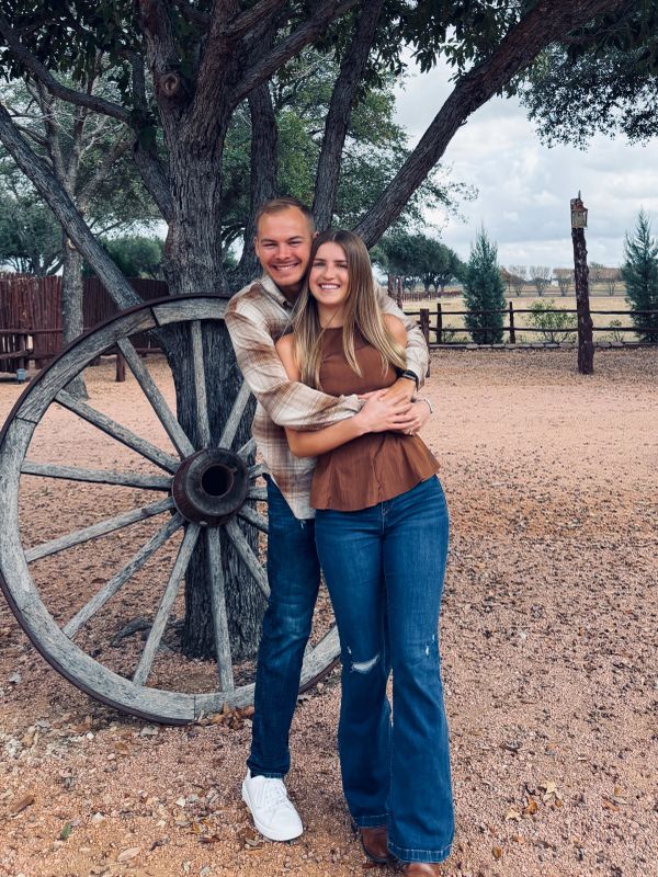 Couple hugging outdoors near a wooden wagon wheel.