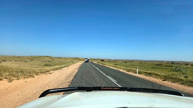 View from a vehicle driving on a straight road through a dry grassland under a clear blue sky.