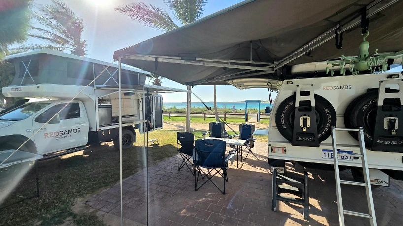 Camping setup with two camper vehicles and chairs under an awning near the beach.