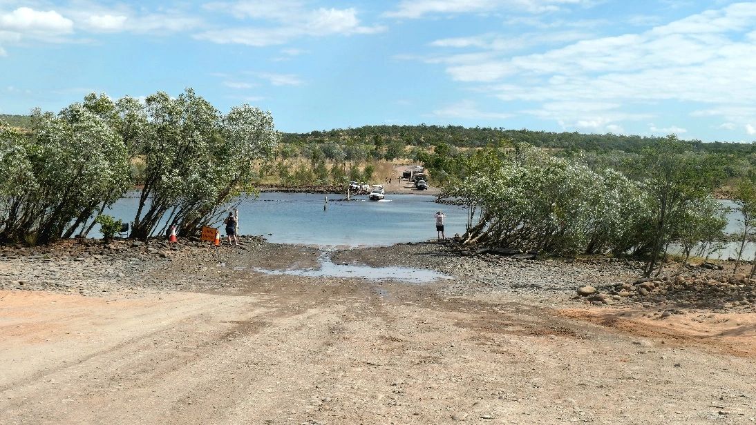 People and vehicles near a shallow river crossing surrounded by trees and rocky terrain.