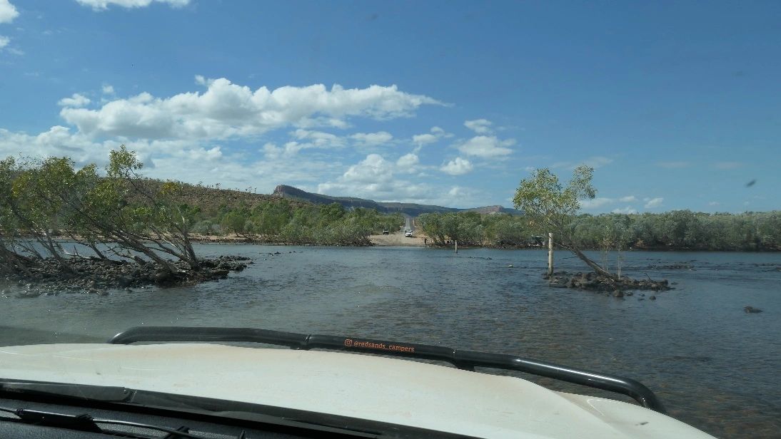 View from a vehicle crossing a shallow river with trees and hills in the background.