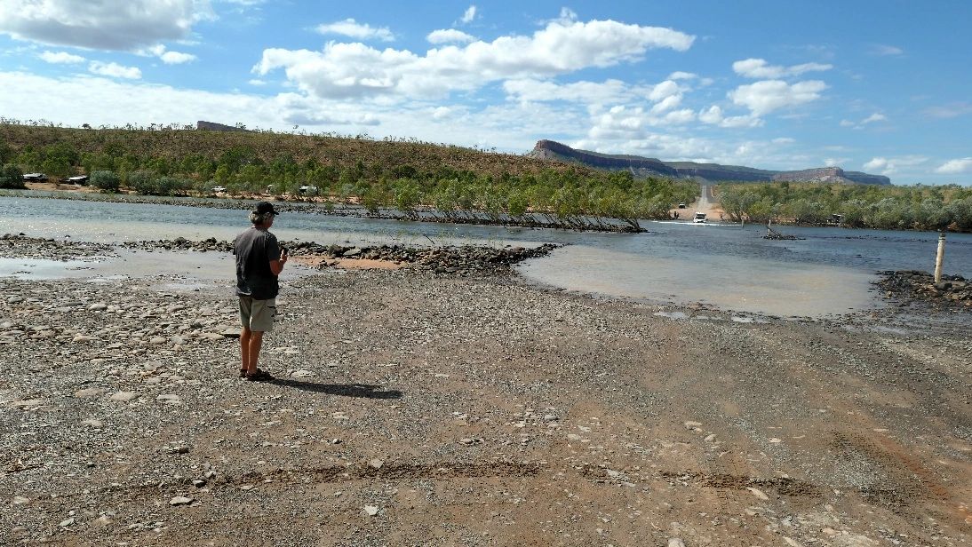 Person stands near a rocky river crossing with trees and hills in the background.