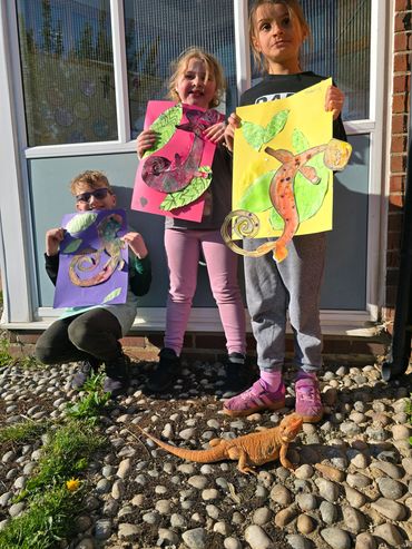 Three kids show colorful lizard drawings with a real lizard on the ground.