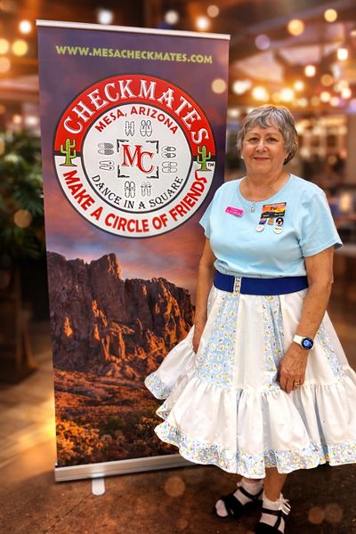 Woman in a square dance outfit stands by a Mesa Checkmates banner.